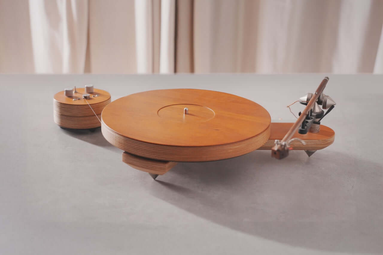 The Arco 40 Plattenspieler, a minimalist wood turntable with separate motor unit and tonearm, sits on a light surface—ideal for vinyl playback—set against beige curtains.