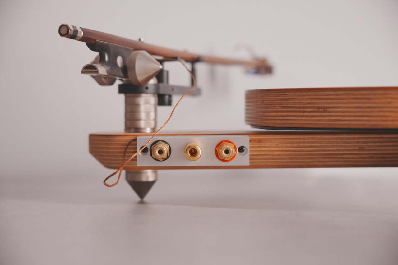 Close-up of the Arco 40 Plattenspieler’s wooden finish, highlighting its tonearm and two gold audio output jacks (one with a red outline) against a neutral background.