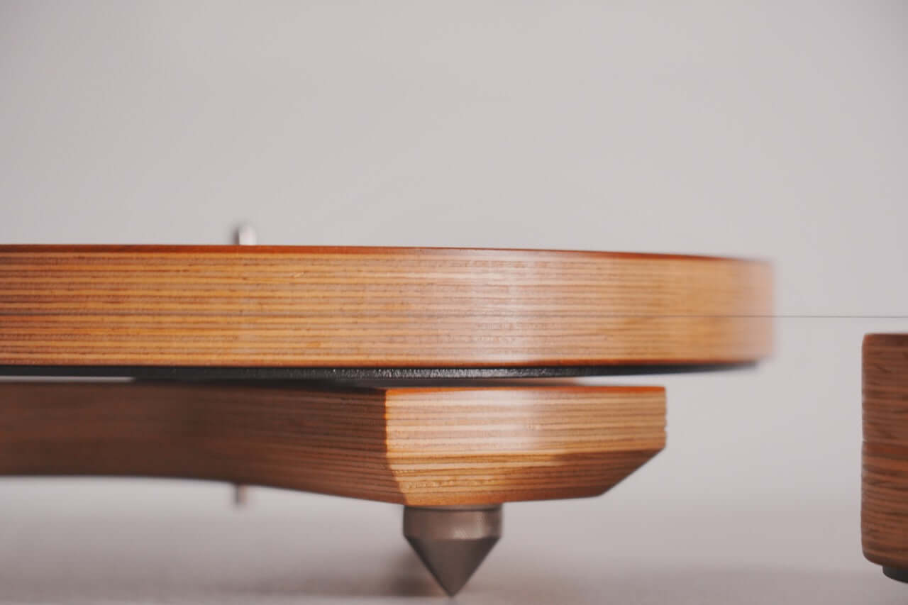 Close-up side view of the Arco 40 Plattenspieler featuring a layered plywood wood base and pointed metal feet, ideal for vinyl enthusiasts, set against a plain light background.
