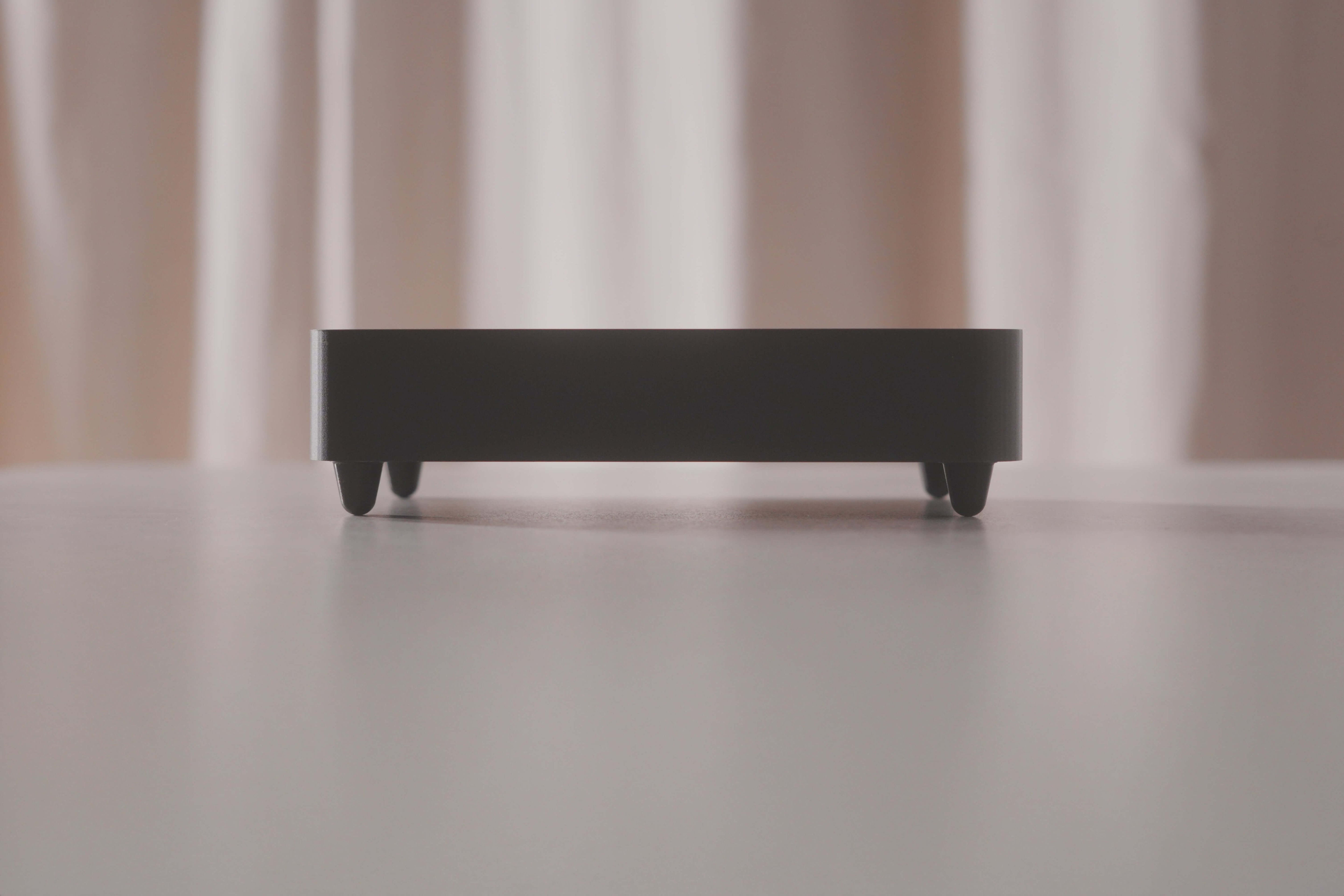 The Schumann Generator S1, a compact black rectangular device with short legs, rests on a smooth light surface and subtly interacts with the Schumann resonance frequency. Beige curtains hang blurred in the background.