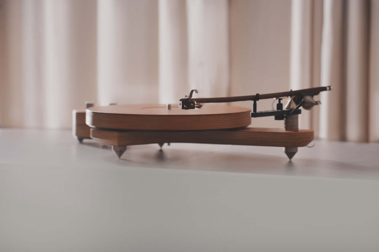 The Arco 40 Plattenspieler, a modern wood turntable with a minimalist design, rests on a smooth surface before light curtains. Its tonearm extends over the circular wooden platter, ready to play your favorite vinyl records.
