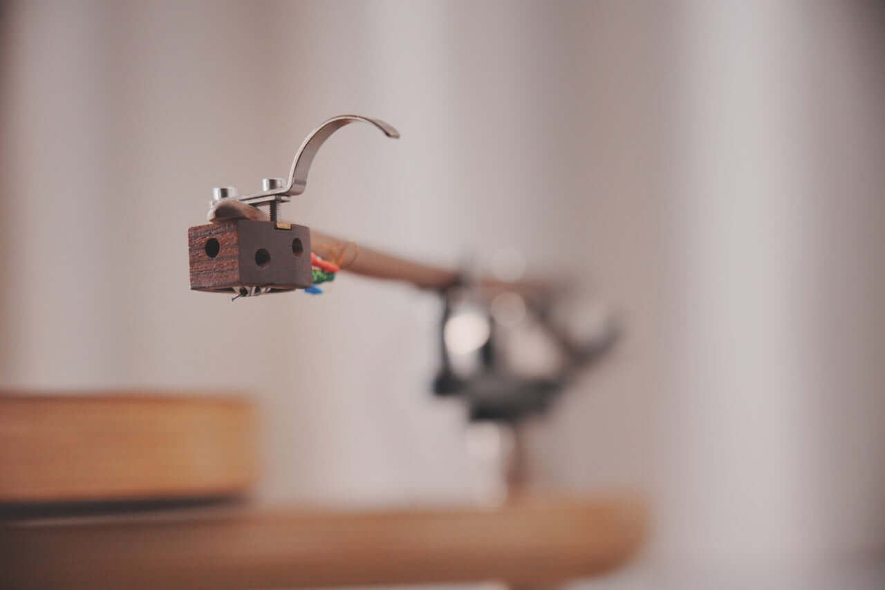 Close-up of the Arco 40 Plattenspieler’s wood tonearm on a vinyl turntable, showing colorful wires and a blurred background. Focus is on the tonearms head, emphasizing its texture and metal components.