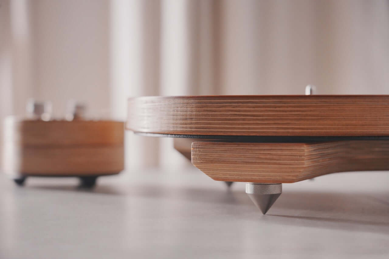 Close-up of the Arco 40 Plattenspieler base, showcasing layered wood and cone-shaped metal feet on a light surface, with a blurred background.