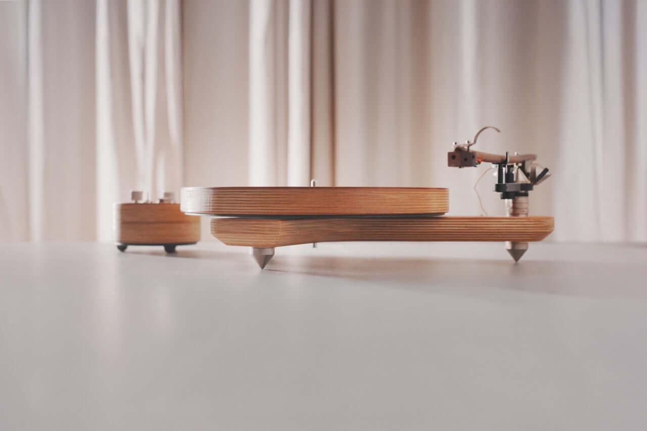 The Arco 40 Plattenspieler, a modern wood turntable with a minimalist design, rests on a light surface. The tonearm and stylus are visible on the right, while soft beige curtains blur in the background, ready for your favorite vinyl.