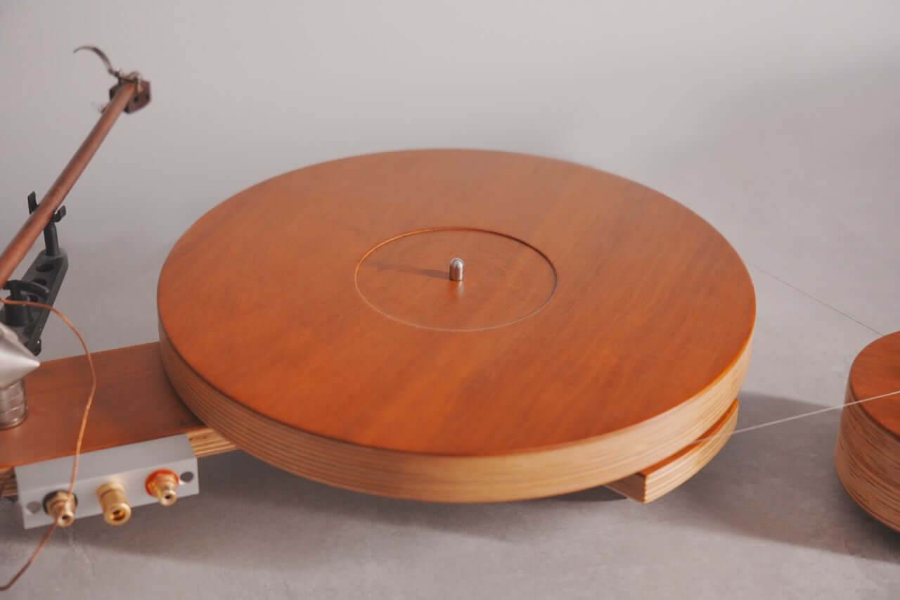 A close-up of the Arco 40 Plattenspieler showcases its circular platter, tonearm, and RCA connectors on a light gray surface. Its minimalistic design with smooth wood grain and metal spindle is ideal for vinyl lovers.