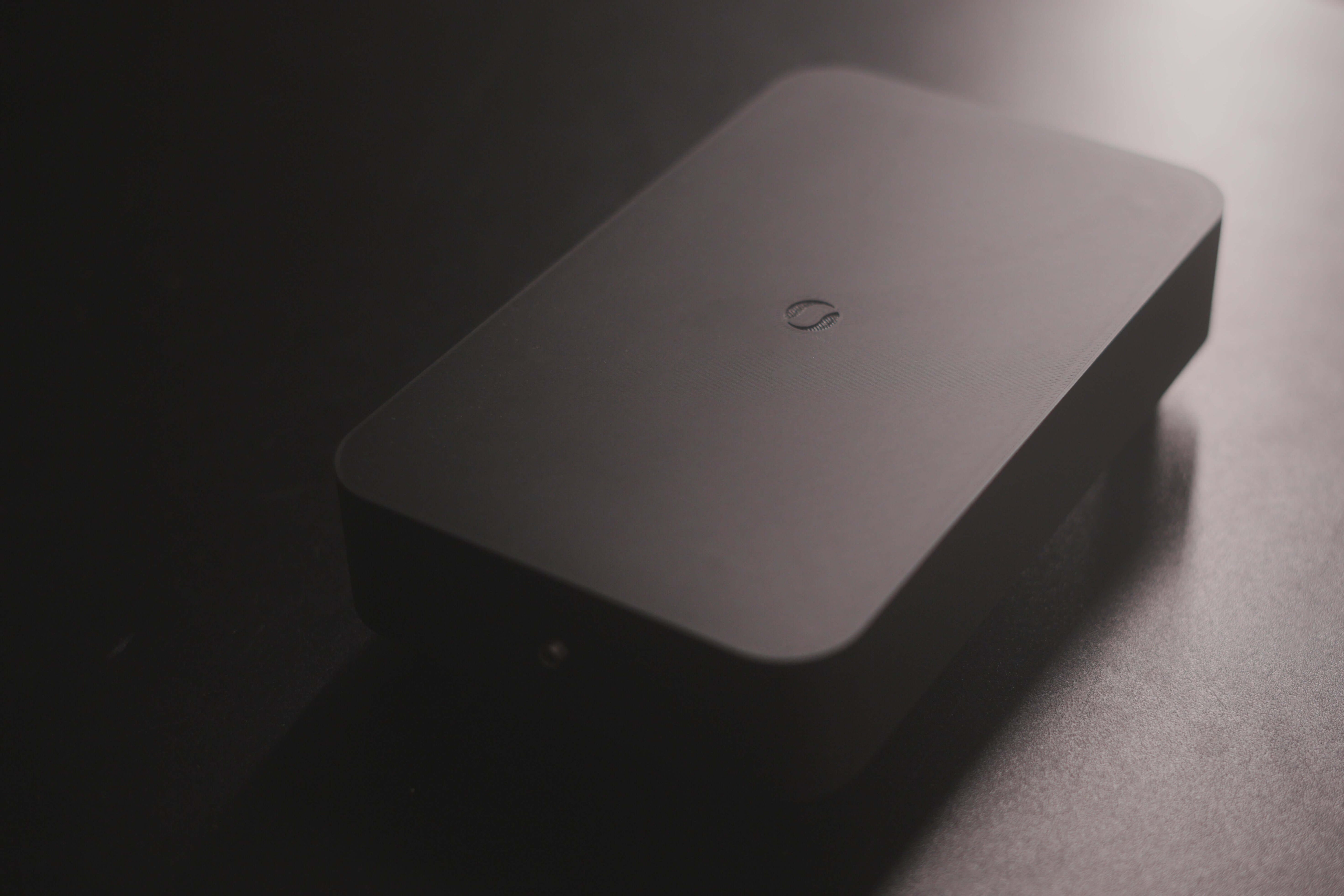 A Schumann Generator S1, a rectangular black device with rounded edges and a small circular symbol on top, sits on a dark surface. Moody lighting highlights its function in modulating das elektromagnetische Feld.